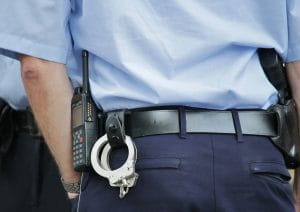 The rear view of a police officer wearing navy blue pants and a sky blue shirt in addition to handcuffs and a communication device on his belt